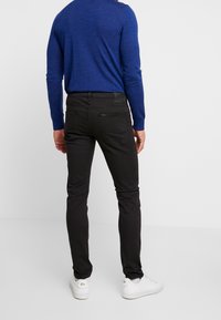 Man som bär svarta slim-fit jeans, blå långärmad skjorta och vita sneakers, står vänd mot en enkel ljusgrå bakgrund.