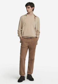 Beige skjorte med knapper over en beige T-shirt med rund hals, parret med brune bukser. Sorte sko fuldender outfittet med et afslappet look.