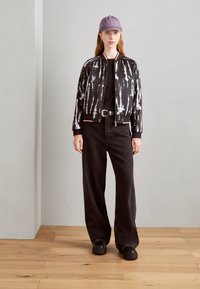 Veste bomber noire tie-dye avec des accents roses, associée à un jean large noir et des chaussures noires chunky. Une casquette violette complète la tenue.