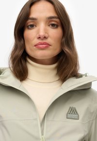 Lichtgroene jacket met ritssluiting en capuchon, met een logo op de borst en een gestructureerde afwerking. G gedragen over een beige coltrui.