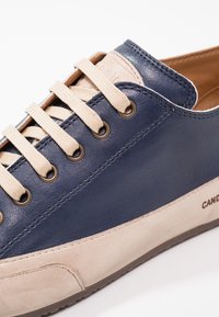 Gros plan d'une basket en cuir bleu marine et beige avec des lacets beiges passés dans des œillets métalliques sur un fond blanc.