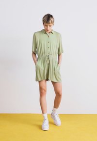 Rompers vert clair à manches courtes avec un devant boutonné, une taille à nouer et des poches. Texture lisse ; stylé avec des baskets blanches et des chaussettes à la cheville.