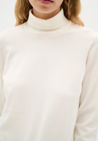 Maglione a collo alto color crema con una texture liscia, caratterizzato da un design aderente e maniche lunghe. Mostra uno stile minimalista e classico.