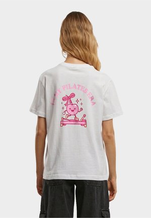 Persona con capelli lunghi che indossa una maglietta bianca con una grafica rosa di un cuore sorridente, che utilizza una macchina per Pilates, con il testo "Nella mia era Pilates."