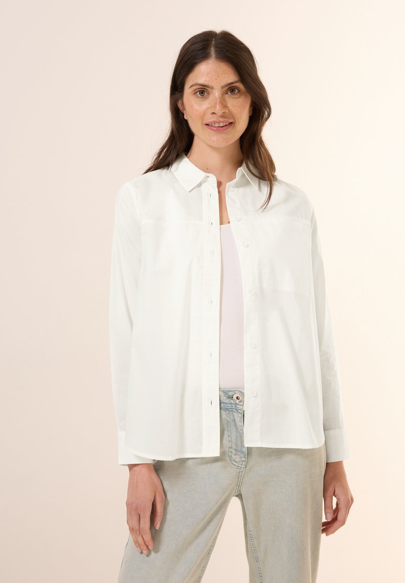 Camicia bianca in cotone, con colletto e maniche lunghe, dalla vestibilità comoda e dettagli di cucitura discreti. Indossata sopra un top leggero.