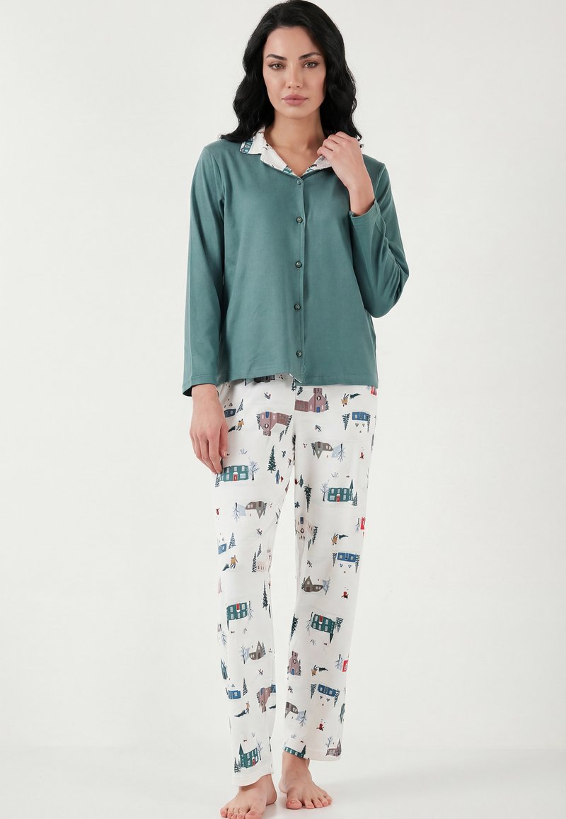 Ženska, oblečena v dolg rokav turkizne barve z gumbi in bele pižamaste hlače s pisanimi vzorci hiš in dreves, stoji bosa na enostavni podlagi.