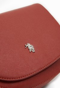 Bolso de mano de cuero texturizado rojo con un acento de logo de caballo plateado, con bordes redondeados y un cierre magnético. Detalle de costura lisa visible.