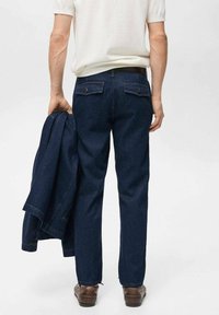 Donkerblauwe denim jeans met twee achterzakken, gedragen met een bijpassende jas, vastgehouden in de linkerhand. Gestructureerd oppervlak, rechte snit.