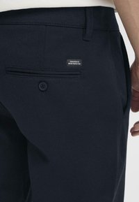 Pantaloni blu navy con texture liscia, caratterizzati da una tasca posteriore, chiusura con bottone e un'etichetta con la scritta "DEDICATED TO DENIM PERFECTION."
