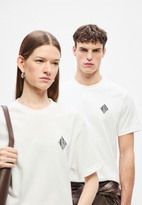 Fehér pólók fekete gyémánt logóval. Az anyag simának tűnik. Az egyik modell egy válltáskát tart a kezében, míg egy másik mögötte áll.