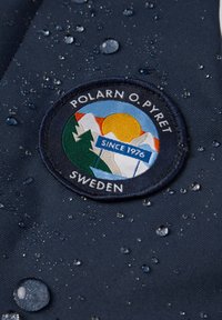 Marine stof met waterdruppels bevat een ronde geborduurde patch. De patch toont bergen, de zon en teksten: "POLARN O. PYRET" en "ZWEEDEN."