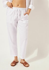 Pantalons en lin blancs avec une taille élastique, des poches obliques et une coupe décontractée. Associés à des sandales à glissière tressées marron.