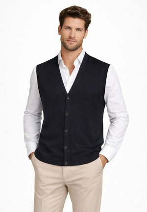 Uomo che indossa un gilet nero con bottoni sopra una camicia bianca e pantaloni beige, in piedi con le mani nelle tasche contro uno sfondo bianco.