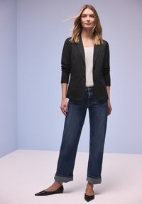 Femme debout portant un jean bleu foncé retroussé, un blazer noir, une chemise blanche et des chaussures plates noires à bouts pointus, sur un fond uni bleu clair.