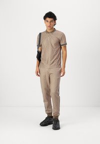 Beige gemustertes Poloshirt und passende Hose mit geometrischem Design, akzentuiert mit schwarz-weißen Streifen an den Seiten. Schwarze Sneaker.