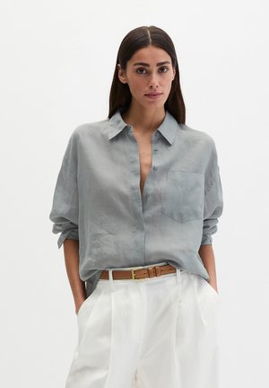 Kobieta o długich ciemnych włosach, ubrana w luźną, jasnoszarą koszulę na guziki, włożoną do białych spodni z wysokim stanem i brązowym paskiem, ręce w kieszeniach.