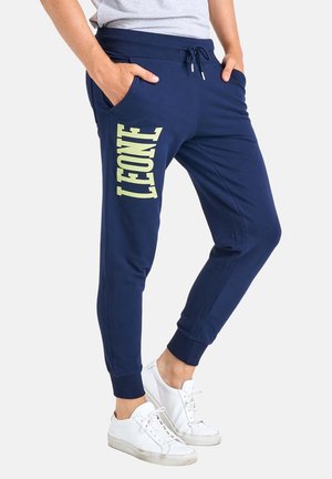Personne portant un pantalon de survêtement bleu marine avec "LEONE" en grandes lettres sur la jambe gauche, associé à des baskets blanches et un haut gris.