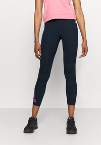 Kvinna i marinblå cropped leggings med rosa logotyp på ena benet och svarta löparskor, stående mot en enfärgad bakgrund.