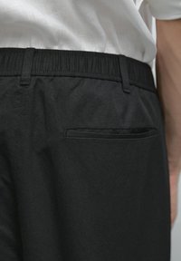 Close-up of back waistband and pocket of black elastic waist pants worn with a tucked white shirt, showing fabric texture and stitching details.