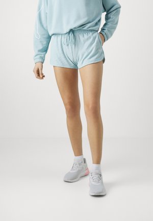 Person iført lyseblå shorts, matchende sweatshirt, hvide sokker og grå sneakers med lyserøde detaljer, stående mod en ensfarvet baggrund.
