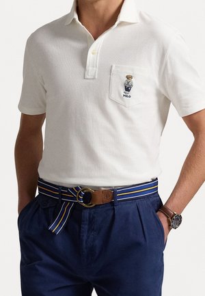 Man draagt een wit poloshirt met berenborduursel op het borstzakje, donkerblauwe broek, gestreepte stoffen riem en polshorloge.
