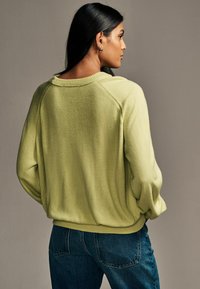 Jasnozielony, oversize'owy sweter o teksturowanym wykończeniu, z prążkowanym wykończeniem i opuszczonymi szwami ramion, zestawiony z ciemnoniebieskimi dżinsami.