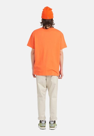 Camiseta de algodón naranja con un corte relajado, combinada con pantalones beige y zapatillas verdes y blancas. El fondo es completamente blanco.