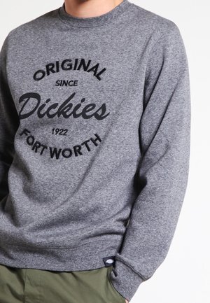 Mężczyzna ubrany w szarą bluzę z czarnym napisem „Original since Dickies 1922 Fort Worth” oraz oliwkowo-zielone spodnie.