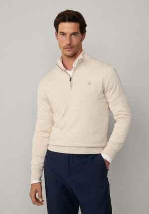 Suéter beige con cremallera y cuello alto, hecho de un tejido suave, presenta puños y dobladillo acanalados, con un logo sutil en el pecho. Se lleva sobre una camisa blanca con cuello.