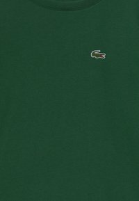 T-shirt en coton vert avec un col rond. Présente un petit logo de crocodile brodé en blanc et rouge sur la poitrine.