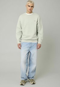 Sweatshirt vert clair avec des poignets et un ourlet côtelés, porté sur une chemise blanche, associé à un jean baggy bleu clair et des baskets grises.