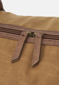 Sac en toile marron avec une fermeture éclair en haut. Comprend deux tirettes en cuir marron et une surface texturée. Détails de couture subtils le long des bords.