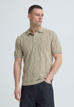 Man met een beige poloshirt met korte mouwen en een golvend breipatroon, en een donkere blauwe spijkerbroek, staand tegen een lichtgrijze achtergrond, kijkend naar rechts.