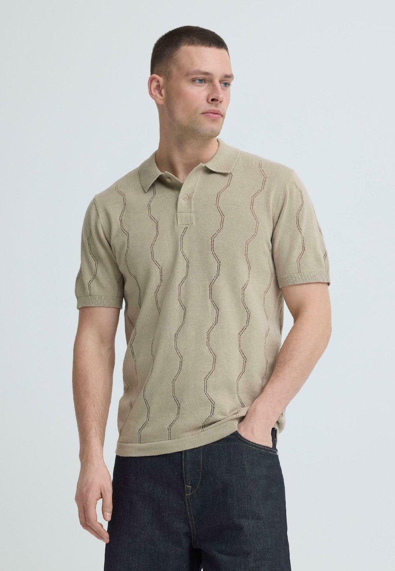 Homme portant un polo beige à manches courtes avec un motif en maille ondulée et un jean bleu foncé, debout devant un fond gris clair, regardant vers la droite.