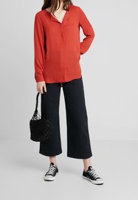 Blouse rouge à manches longues avec un décolleté en V, associée à un pantalon noir à jambes larges et des baskets noires. Accessoire : un petit sac tressé noir.