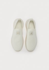 Bijele slip-on cipele s teksturiranim pletenim gornjim dijelom, mekim elastičnim stranama i malom logotipnom oznakom na jeziku. Sadrži lagani potplat.