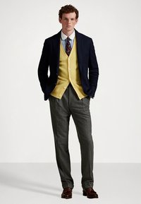 Marineblå blazer over en gul cardigan, hvit skjorte og mønstret slips. Grå bukser med teksturert finish. Brune sko fullfører antrekket.