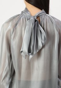 Blusa di seta grigia chiara con chiusura a fiocco sul retro. Caratterizzata da un colletto plissettato e una trama trasparente, crea una forma morbida e fluida.