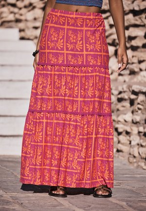 Kvinde iført en lang lyserød nederdel med orange blomstrede firkantede mønstre, sorte sandaler og en blå cropped top, stående udendørs på stenetrin.