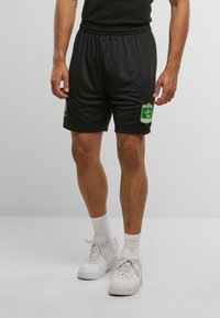 Man wearing black sports shorts with a green and yellow "Fussballclub Kitzbühel" logo, white socks, and white sneakers standing on a white floor.
