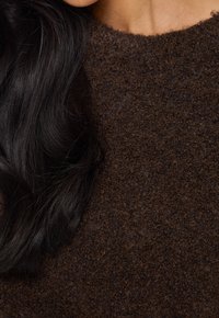 Brun ulltröja med en texturerad, tjock stickad design, med en rund halsringning, som ligger bredvid långt, rakt mörkt hår.