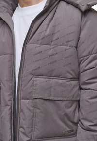 Chaqueta acolchada gris con cierre de cremallera, gran bolsillo de parche y patrón de logo "PGDR" repetido en la tela. Diseño texturizado y acolchado.