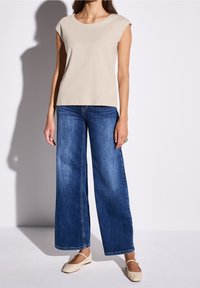 Top beige a manica corta abbinato a jeans in denim blu a gamba larga. L'outfit presenta un design semplice, una texture liscia e uno stile casual classico.