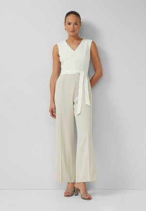 Woman wearing sleeveless white wrap top and high-waisted wide-leg beige trousers with heeled sandals, standing against a plain light wall.