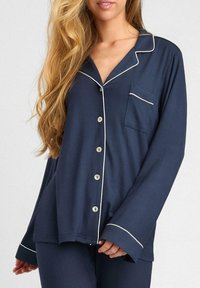 Granatowy top piżamy z długimi rękawami, perłowymi guzikami i białymi wykończeniami. Posiada kieszeń na piersi oraz luźny krój. Miękka tekstura.