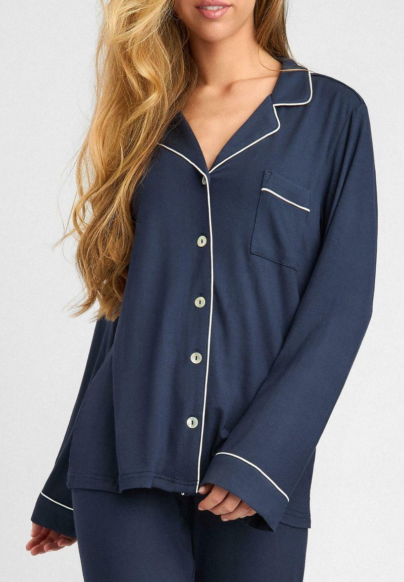 Granatowy top piżamy z długimi rękawami, perłowymi guzikami i białymi wykończeniami. Posiada kieszeń na piersi oraz luźny krój. Miękka tekstura.