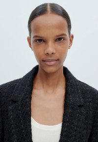 Zwarte geweven blazer met een gestructureerd patroon, voorzien van notched lapels. Daaronder een goed aansluitende witte geribde tanktop. Eenvoudig, strak ontwerp.