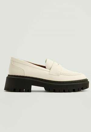 Mocasines de cuero crema con punta redondeada, con una suela negra gruesa y con ranuras, y una correa cruzando el empeine; detalles mínimos de costura.