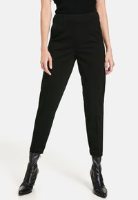 Pantalon noir sur mesure à taille haute, doté de détails plissés et d'une coupe fuselée. Le tissu a une texture lisse; porté avec des bottines noires.