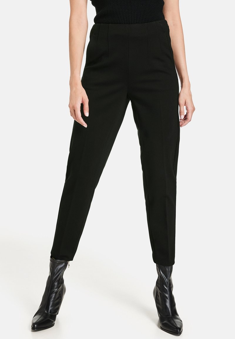 Pantalon noir sur mesure à taille haute, doté de détails plissés et d'une coupe fuselée. Le tissu a une texture lisse; porté avec des bottines noires.